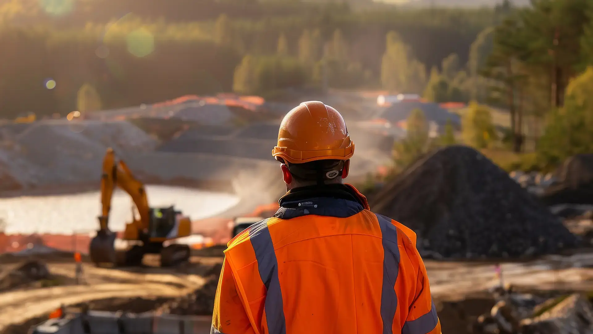 Rear view of a construction worker in an orange safety jacket and hard hat overlooking an active mining or excavation site at sunset.