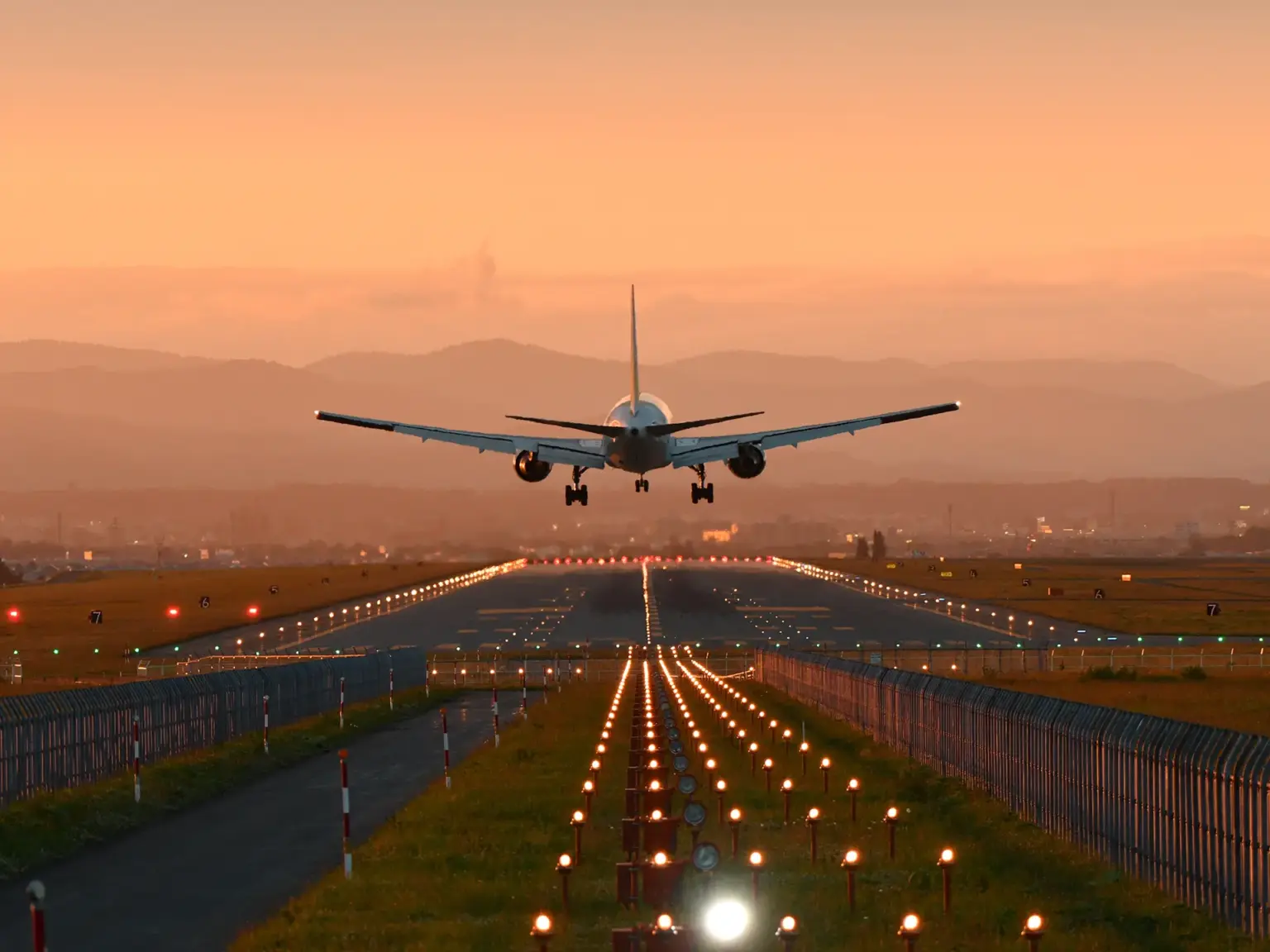Rear view of a commercial aircraft descending toward a runway at sunset, with runway lights glowing and mountains in the distance.