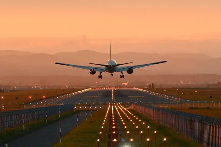 Rear view of a commercial aircraft descending toward a runway at sunset, with runway lights glowing and mountains in the distance.