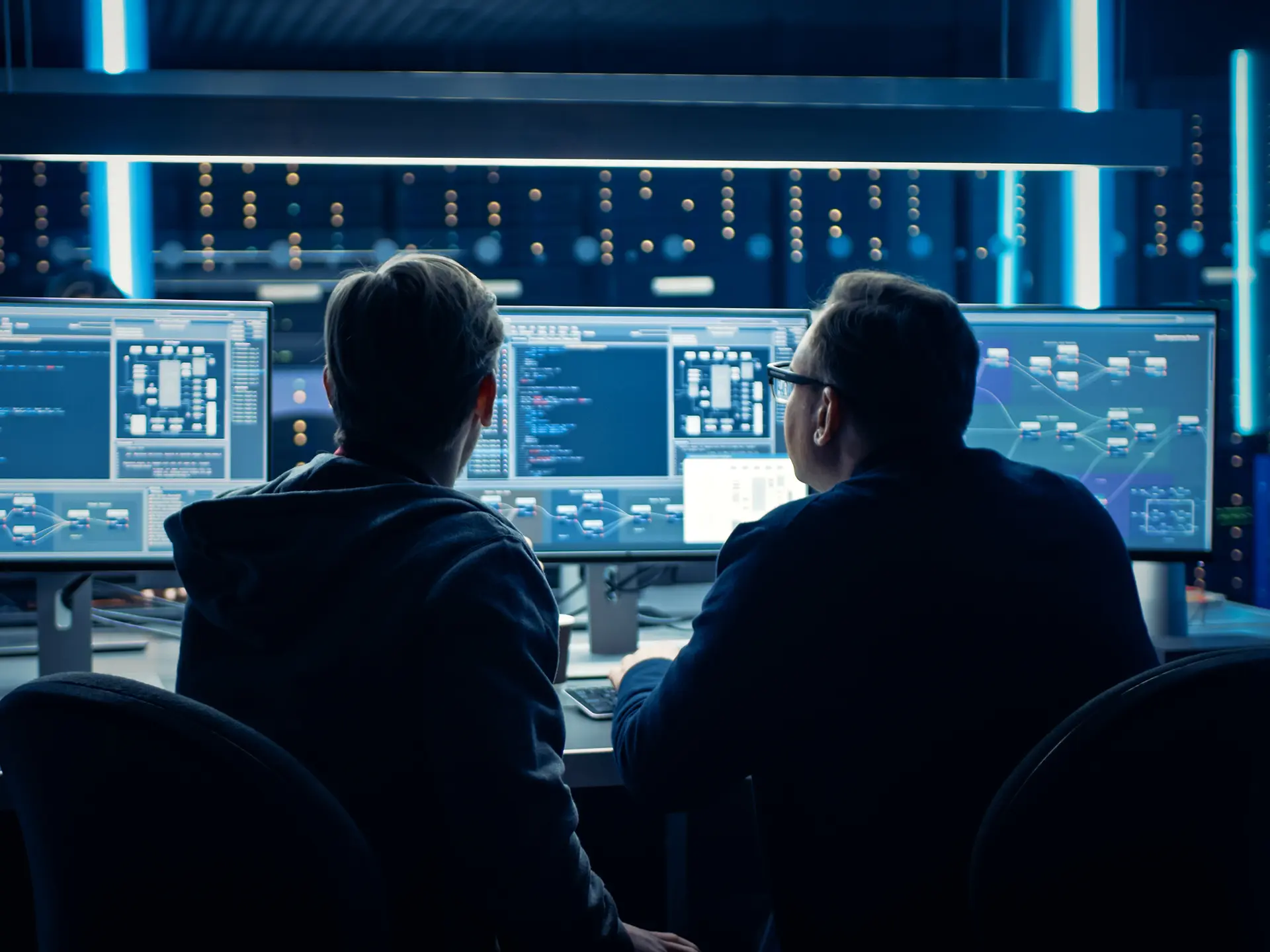 Two people seated at a desk in a data center, looking at multiple large monitors displaying code and system diagrams.