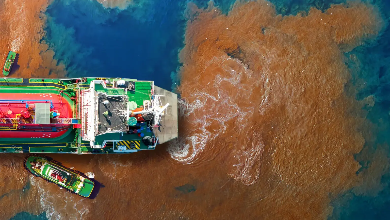 Aerial view of a large vessel surrounded by a wide plume of brown, polluted water spreading into blue sea.