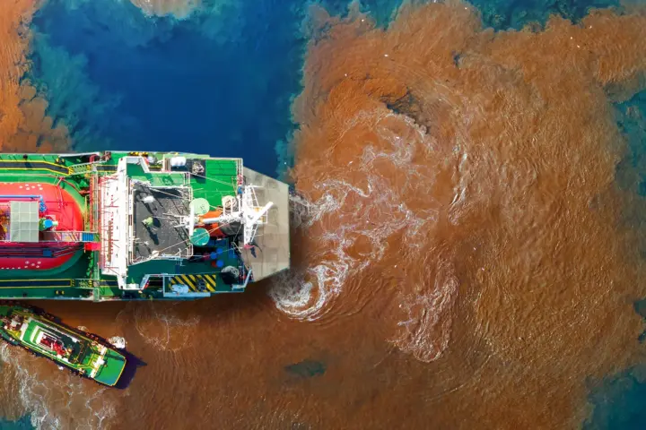 Aerial view of a large vessel surrounded by a wide plume of brown, polluted water spreading into blue sea.