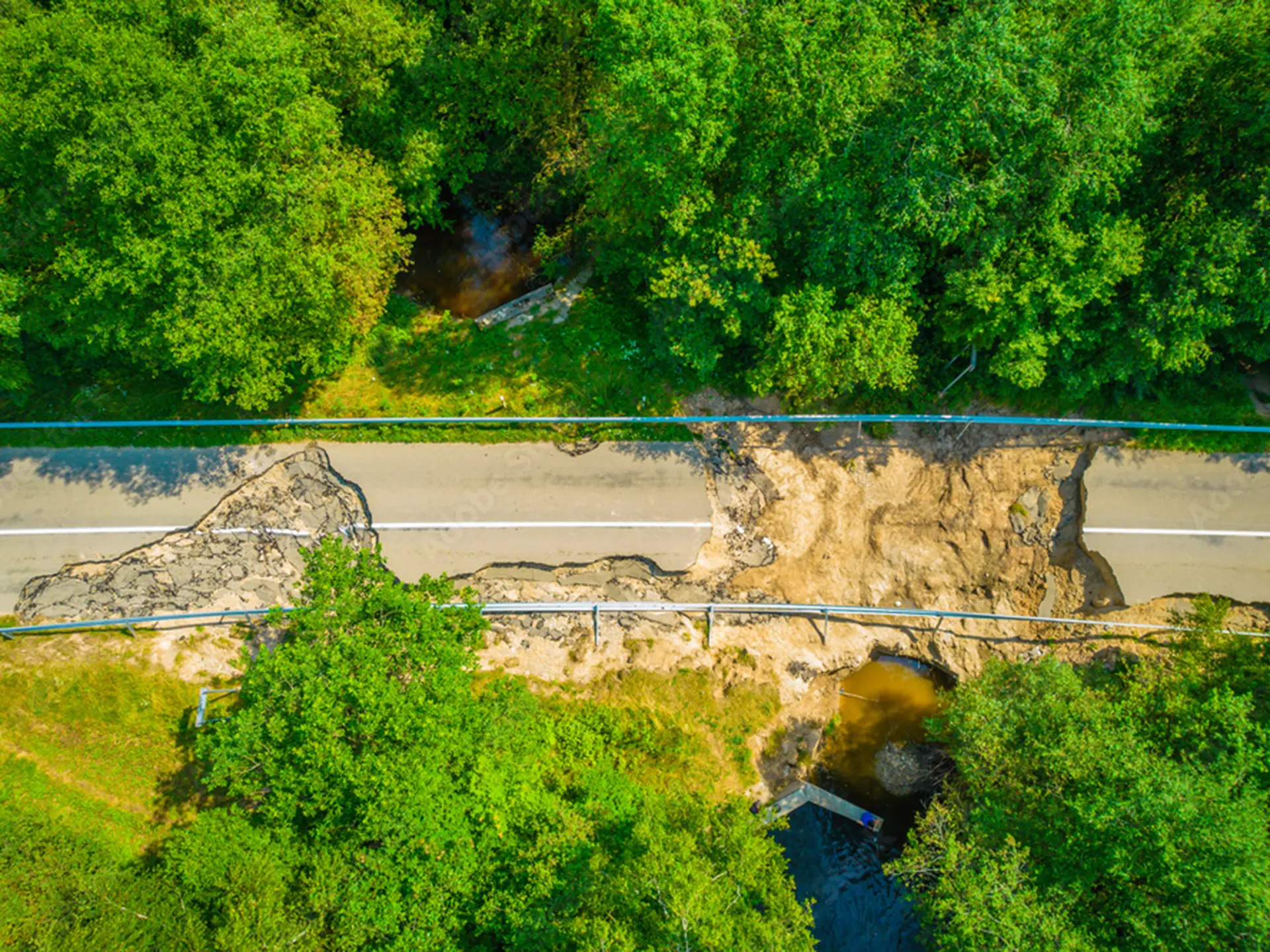 Aerial view of a damaged road with a large section washed away, exposing soil and water beneath, surrounded by green vegetation.