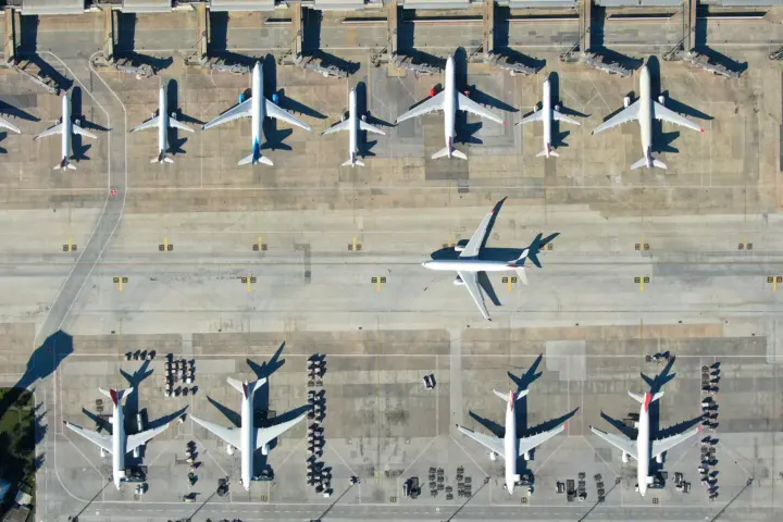 Top-down aerial view of multiple commercial airplanes parked at airport gates, with one aircraft taxiing along the runway.